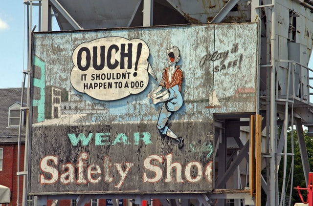 Faded billboard of cartoon man holding foot in pain; text: Ouch! It shouldn't happen to a dog. Play it safe! Wear safety shoes