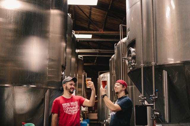 Two brewery employees, standing between rows of large chilled tanks, examining freshly poured pints of beer