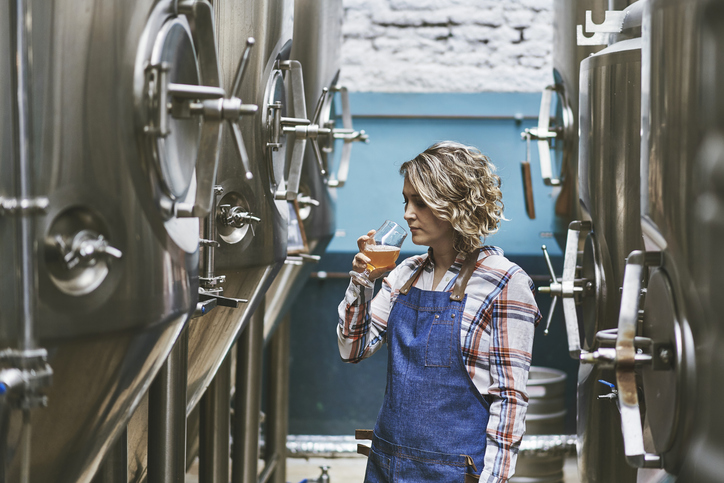 Female craft brewer standing between vats and examining beer sample in glass.