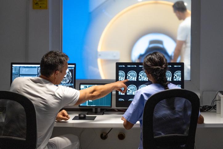 Rear view of male and female MRI technologists sitting at the console in the operating room and operating the MRI scanner